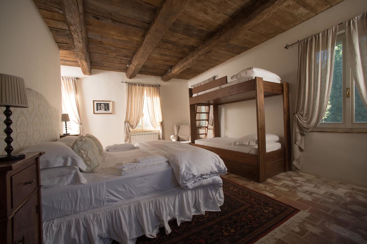 A bedroom with a wooden bunk bed, a single bed, a bedside table with a lamp, and a window with curtains.