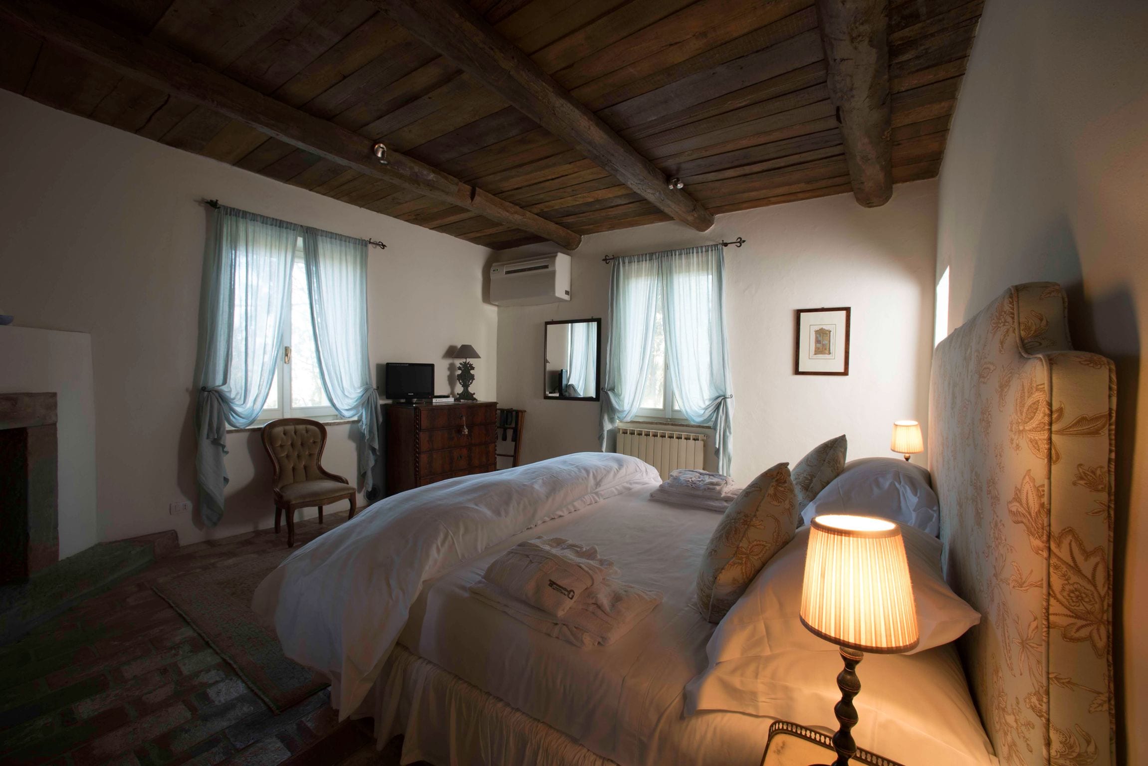 A bedroom with a wooden ceiling, a bed with white linens, a bedside lamp, a chair, a dresser, and a fireplace.