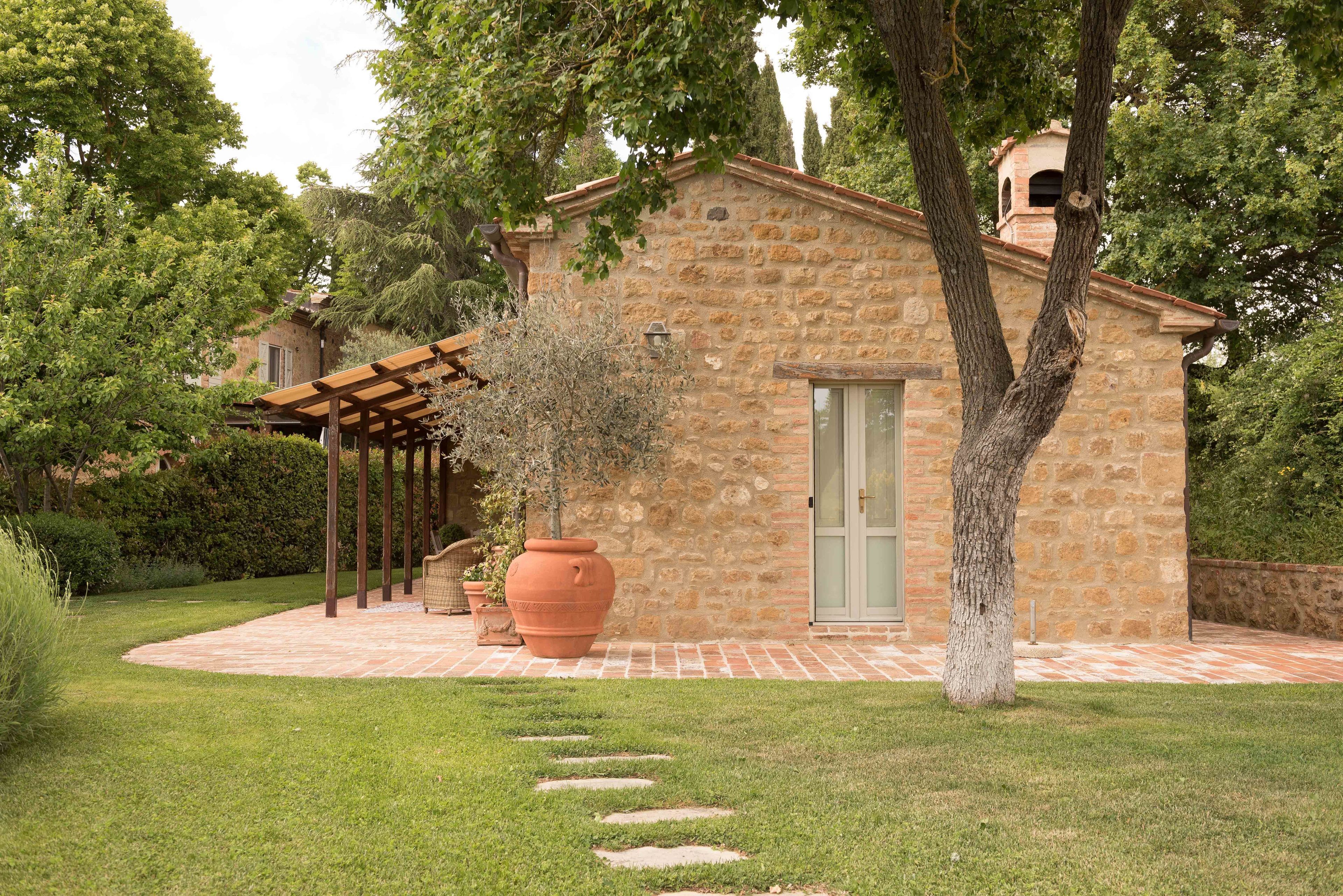 A stone building with a wooden pergola, a large terracotta pot, and a paved pathway.