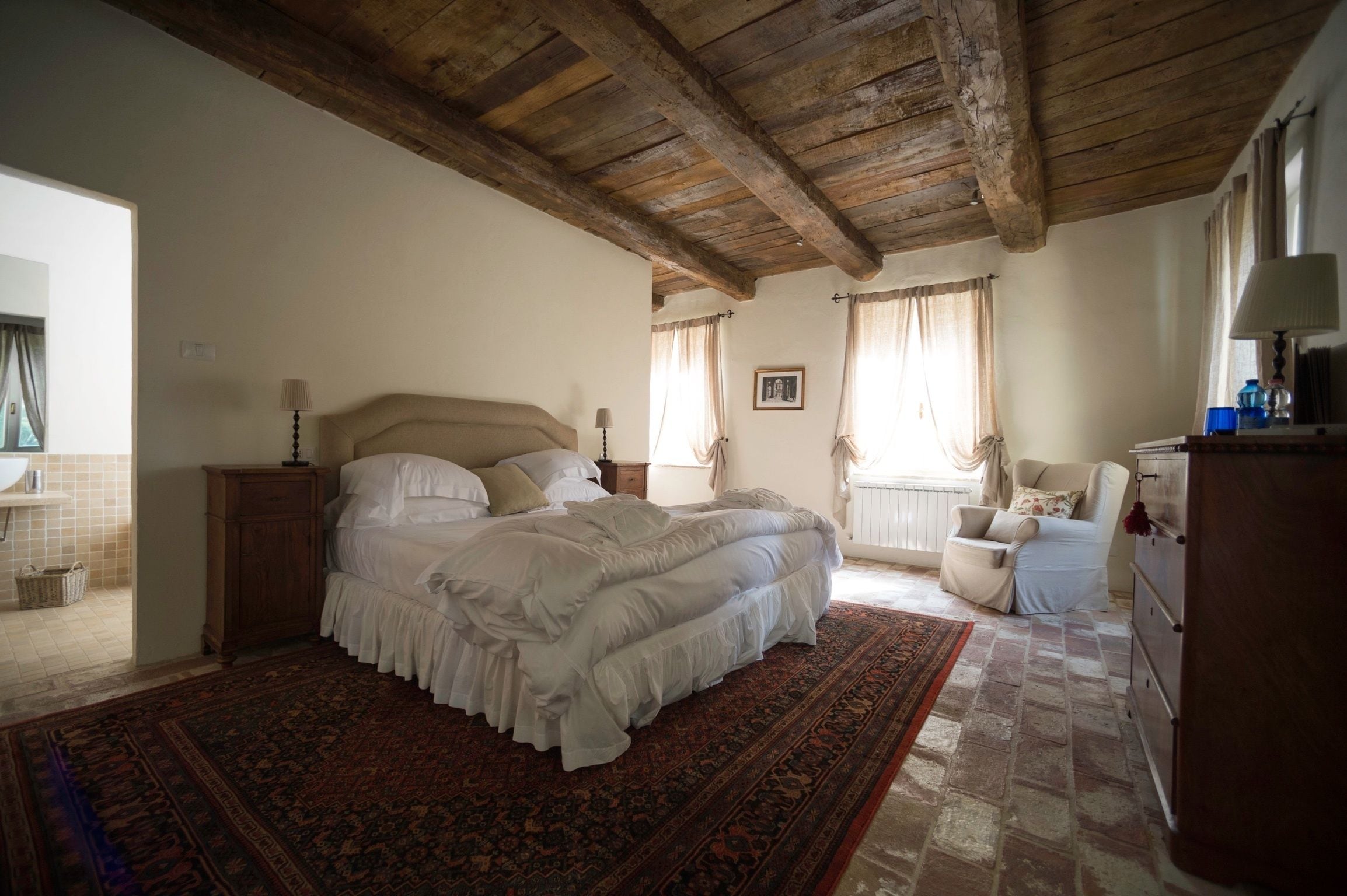 Un dormitori amb un llit gran, bigues de sostre de fusta, una petita cadira, un taulell i una finestra amb cortines.