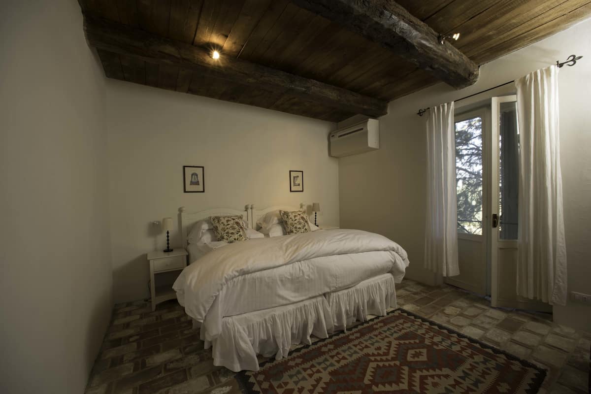A bedroom with a wooden ceiling, a bed with white bedding, a patterned rug, and a window with curtains.