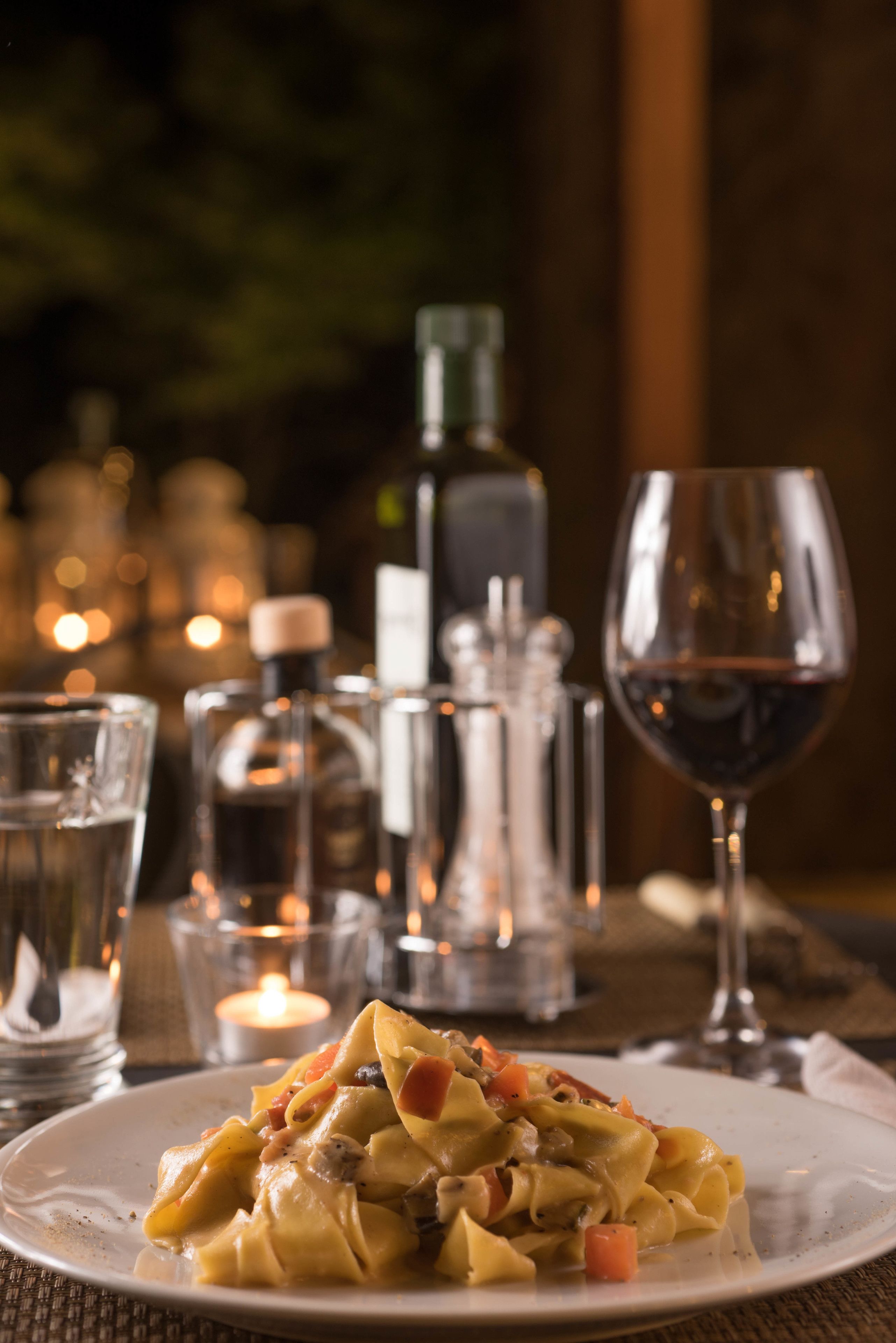 A plate of pasta with vegetables, a glass of red wine, and a candle on a table with salt and pepper shakers in the background.