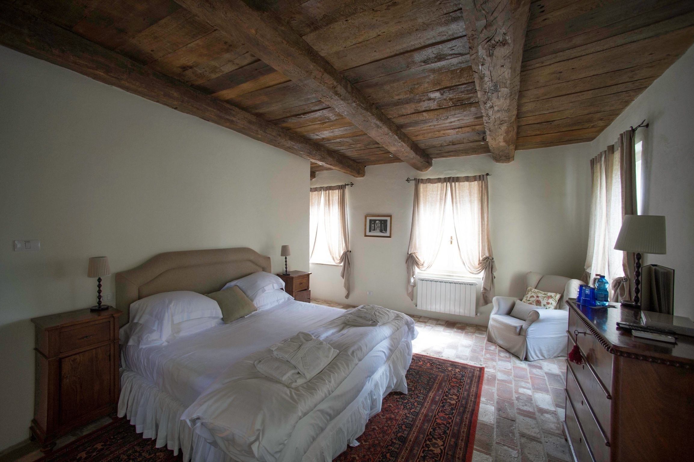 A bedroom with a large bed, wooden ceiling beams, a radiator, a chair, and a dresser.