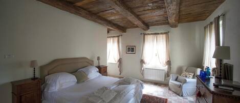 Quarto com uma cama grande, vigas de madeira no teto, radiador, cadeira e cômoda.