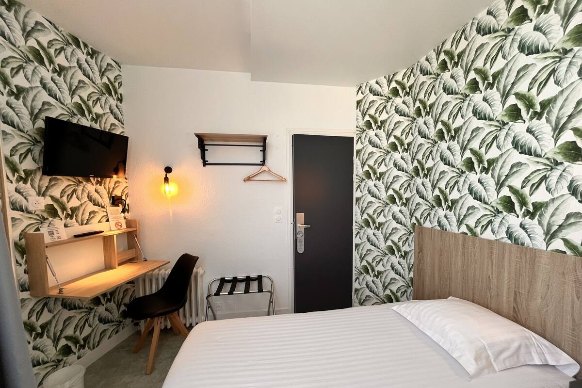Ein Hotelzimmer mit Bett, Schreibtisch mit Stuhl, Fernseher, Wandleuchte und einer Tür.