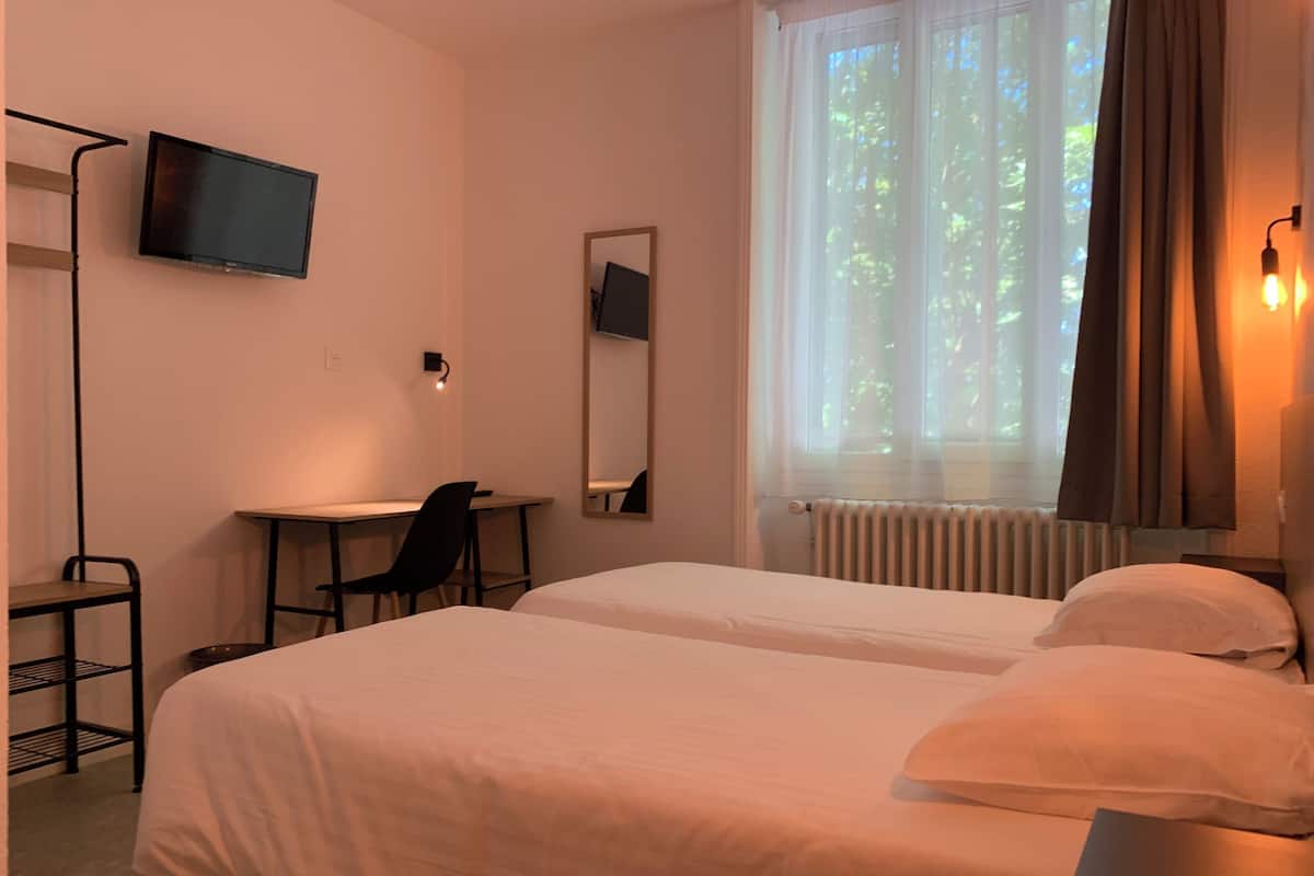 Ein Hotelzimmer mit zwei Betten, einem Schreibtisch, einem Stuhl, einem Fernseher, einem Spiegel und einem Fenster mit Vorhängen.
