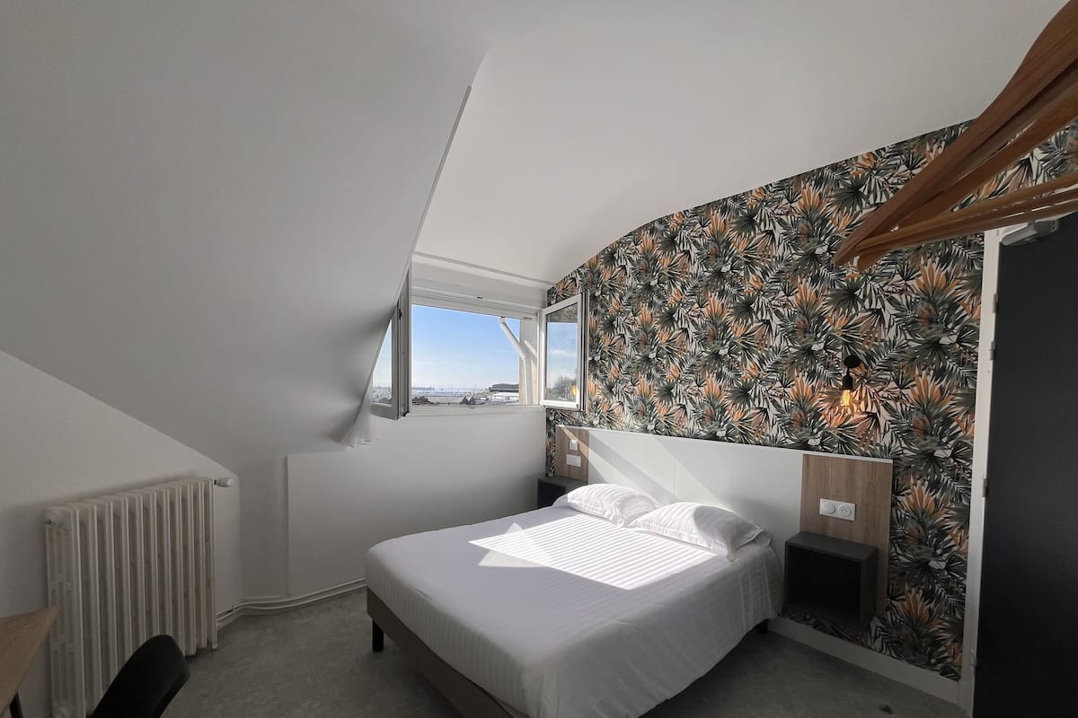 Ein Schlafzimmer mit Bett, Fenster, Heizkörper und einer Wand mit Blätermotiv.