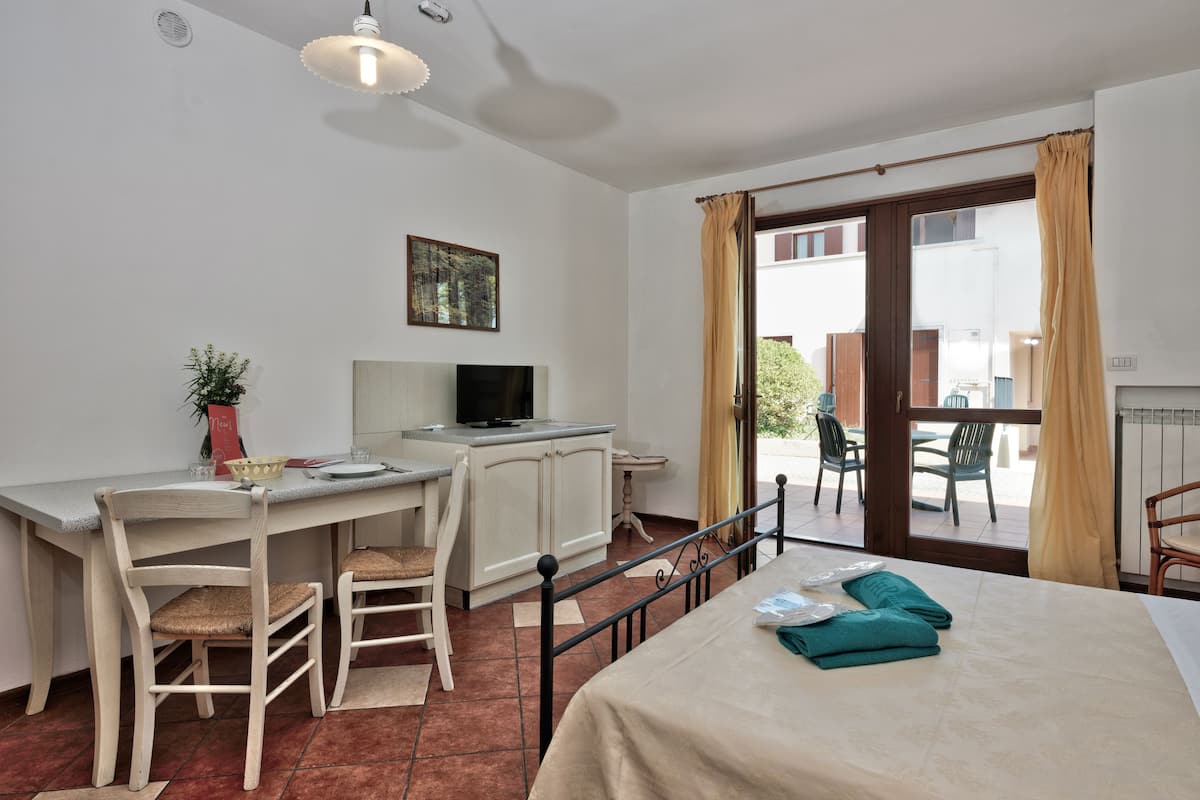 A room with a bed, a desk with a computer, a chair, a small table with a plant, and a window with curtains.