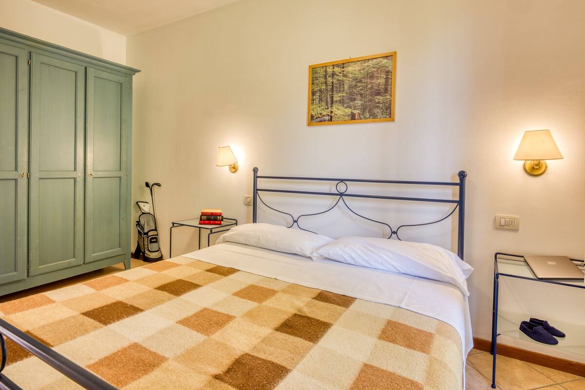 A bedroom with a checkered carpet, a metal bed frame, a bedside table with a lamp, a framed picture on the wall, and a green wardrobe.