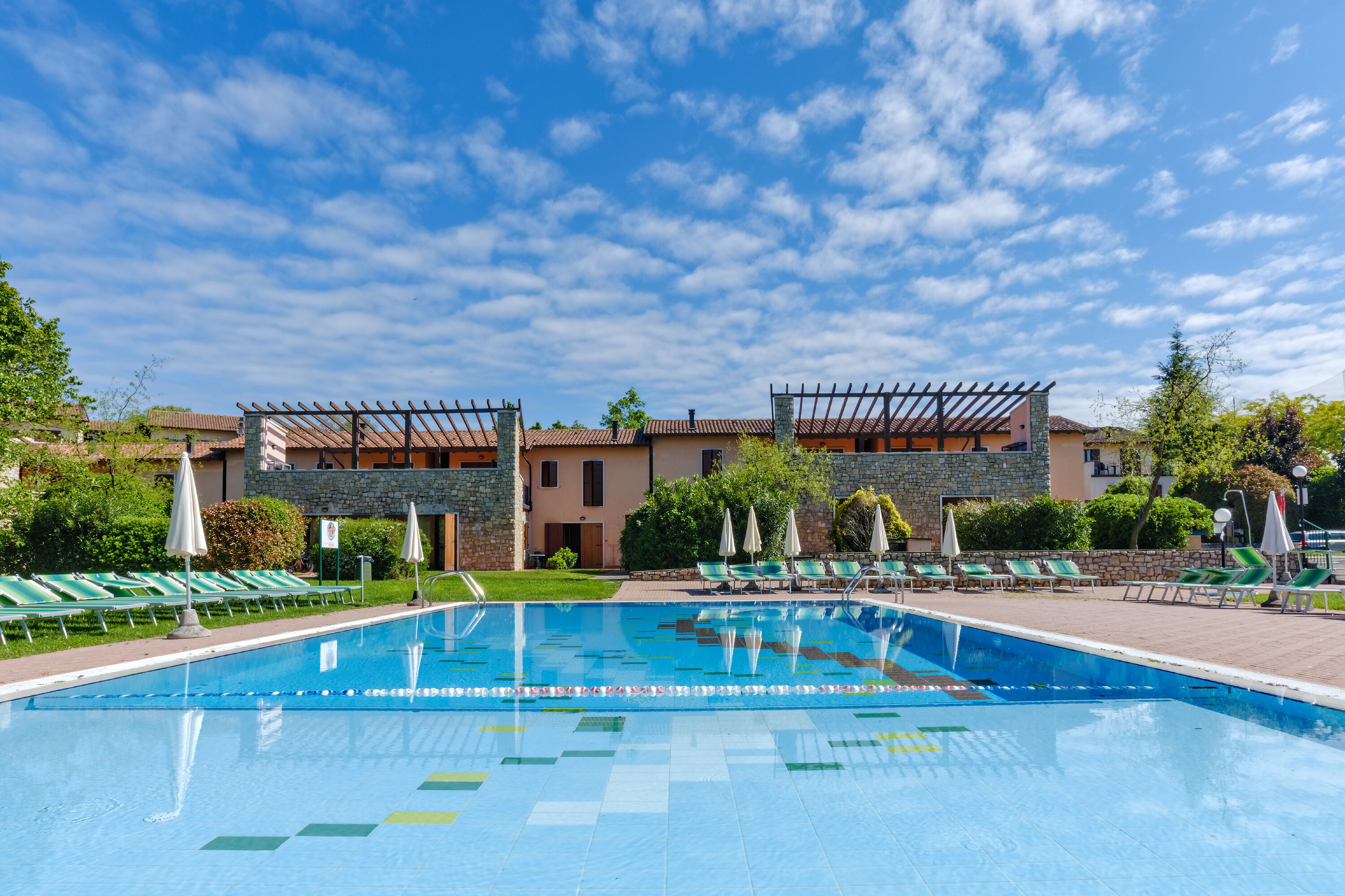 Una piscina con sillas de descanso y sombrillas, rodeada de edificios con fachadas de piedra y estuco.