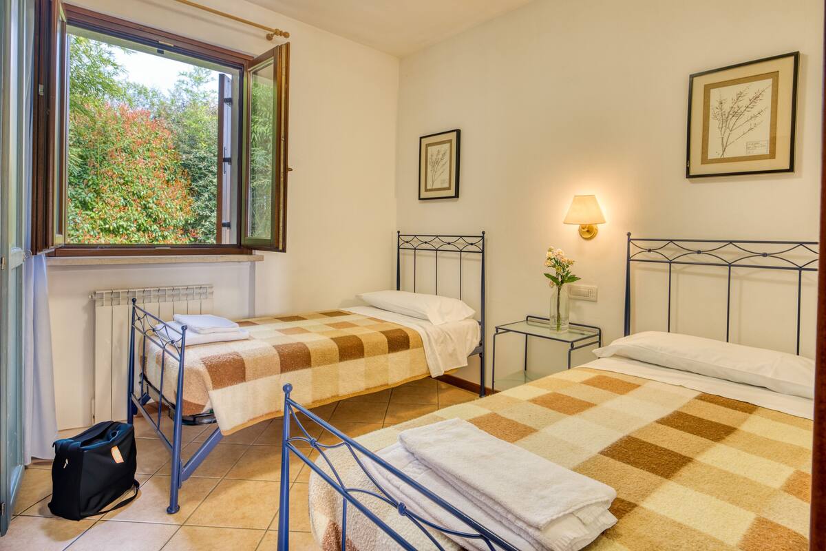 A room with two beds, a window with an open shutter, a radiator, a bedside table with a lamp, and a framed picture on the wall.