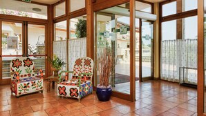 Un hall con puertas de vidrio con marco de madera, dos sillas coloridas, una planta en maceta y un piso de baldosas.
