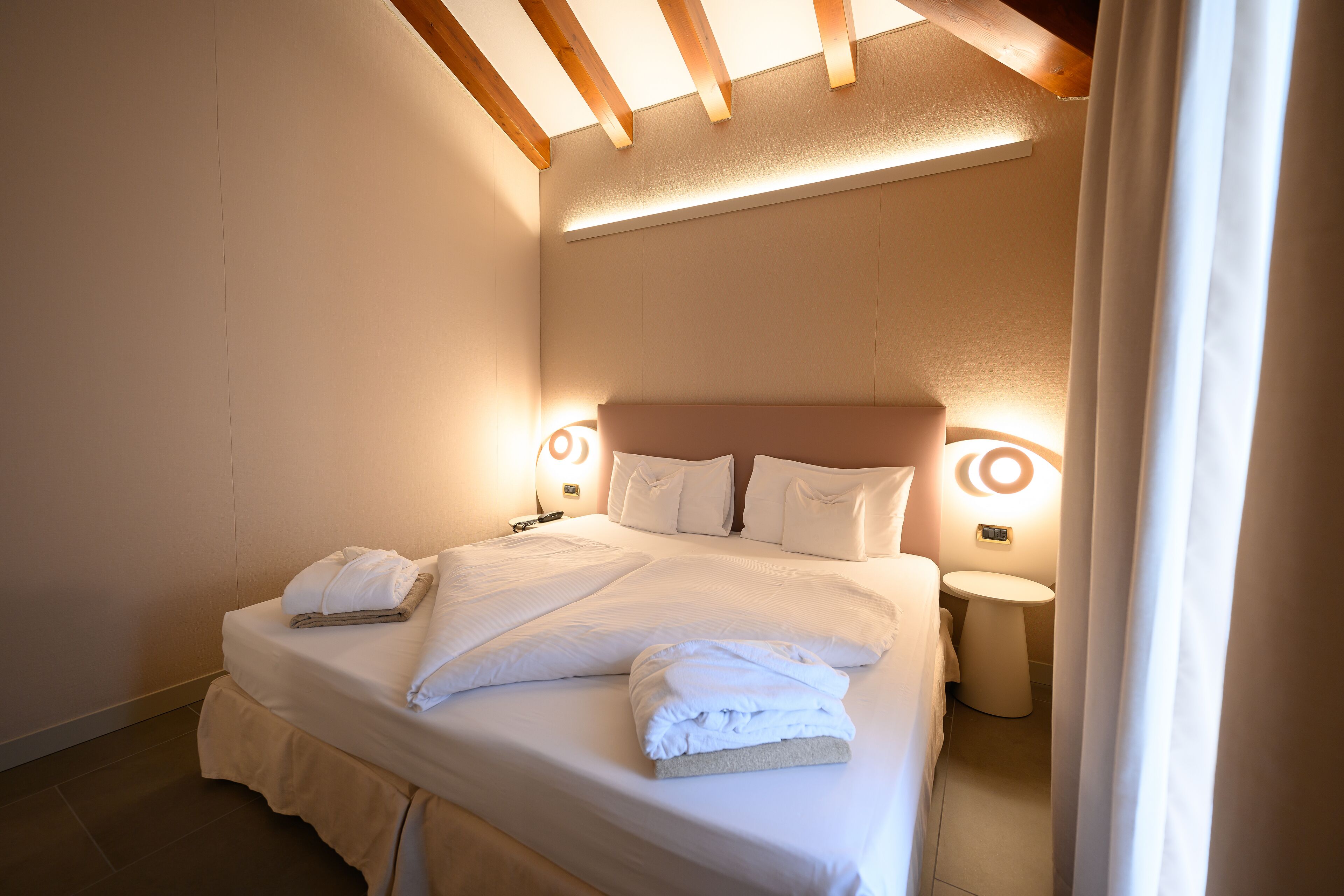 A neatly made bed with white linens and towels, a bedside table with a lamp, and a small bedside table with a clock.