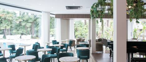 A modern indoor space with round tables and teal chairs, large windows with a view of palm trees, and a piano.