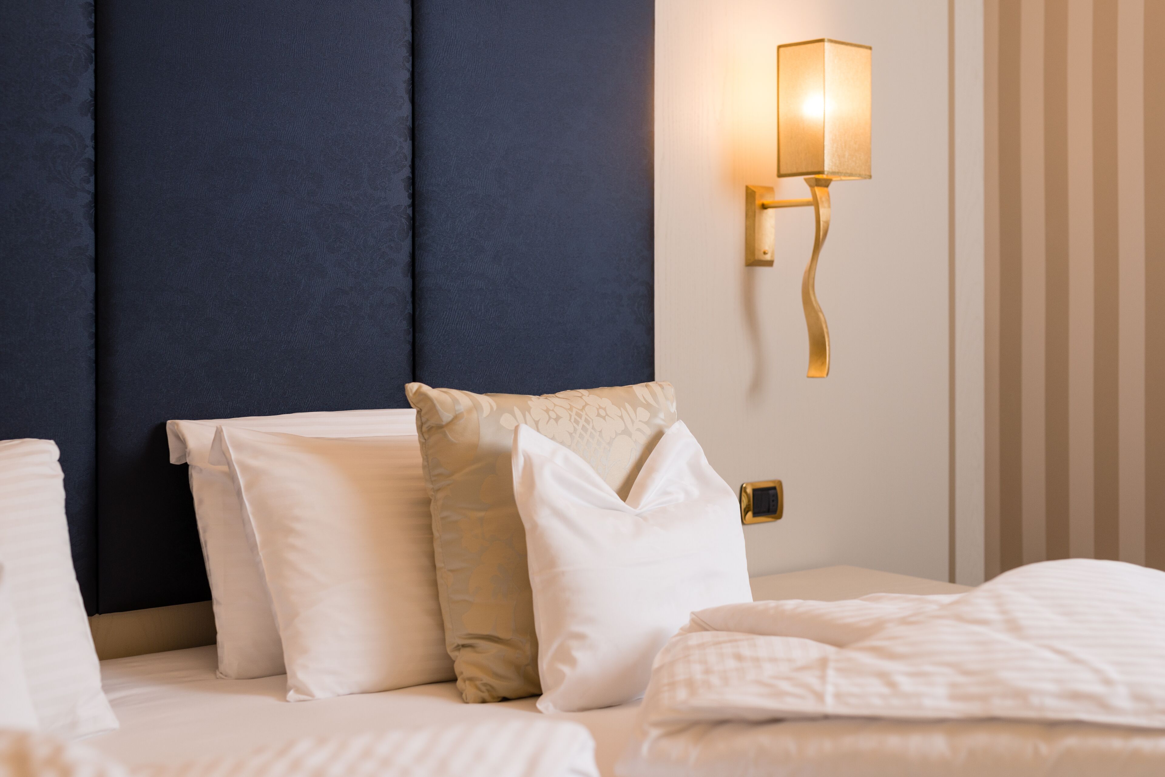 Ein ordentlich bezogenes Bett mit weißer Bettwäsche und mehreren Kissen, ein dunkelblaues Kopfbrett und eine wandmontierte Lampe.