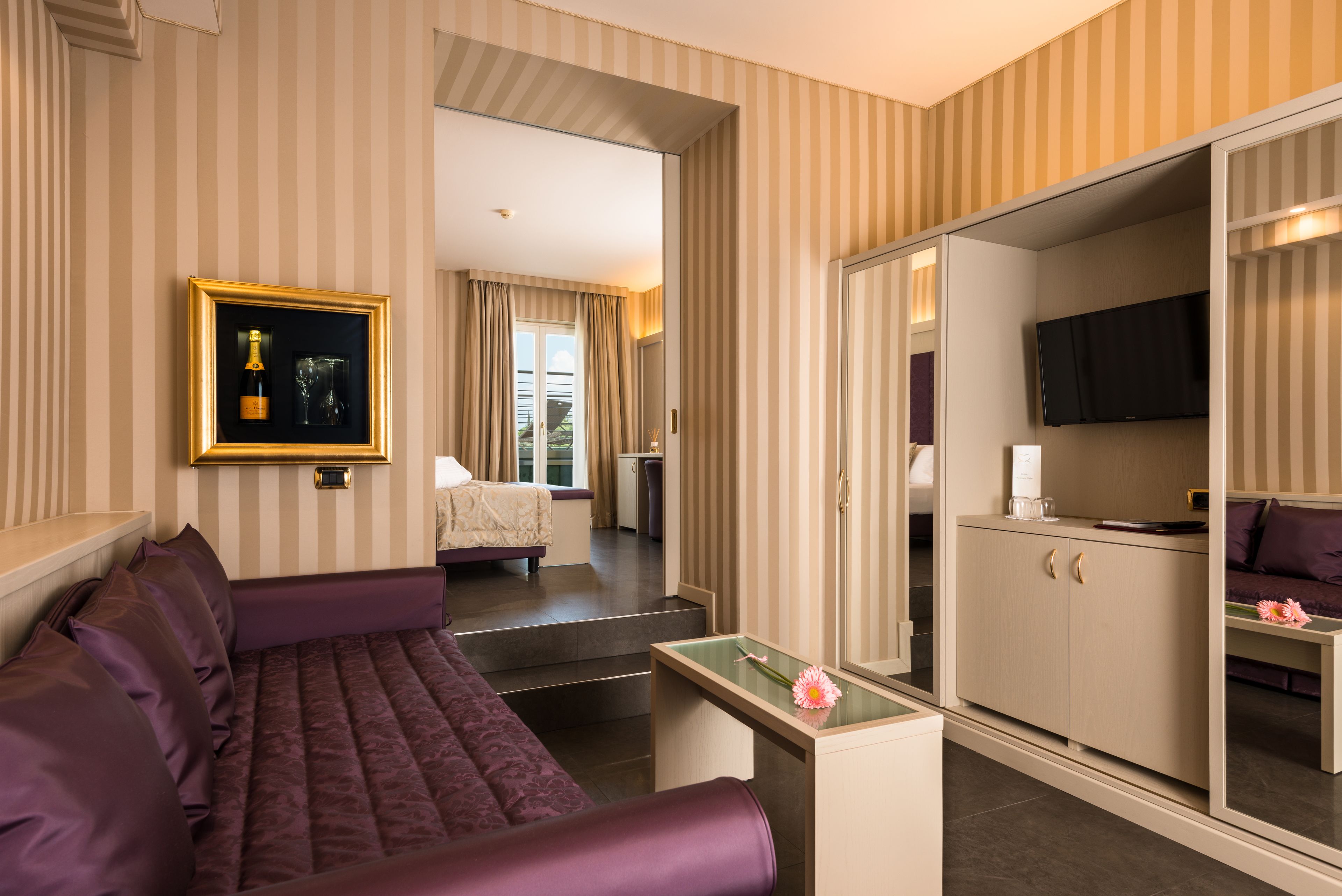 A hotel room with a bed, a sofa, a TV, a minibar, and a mirror.