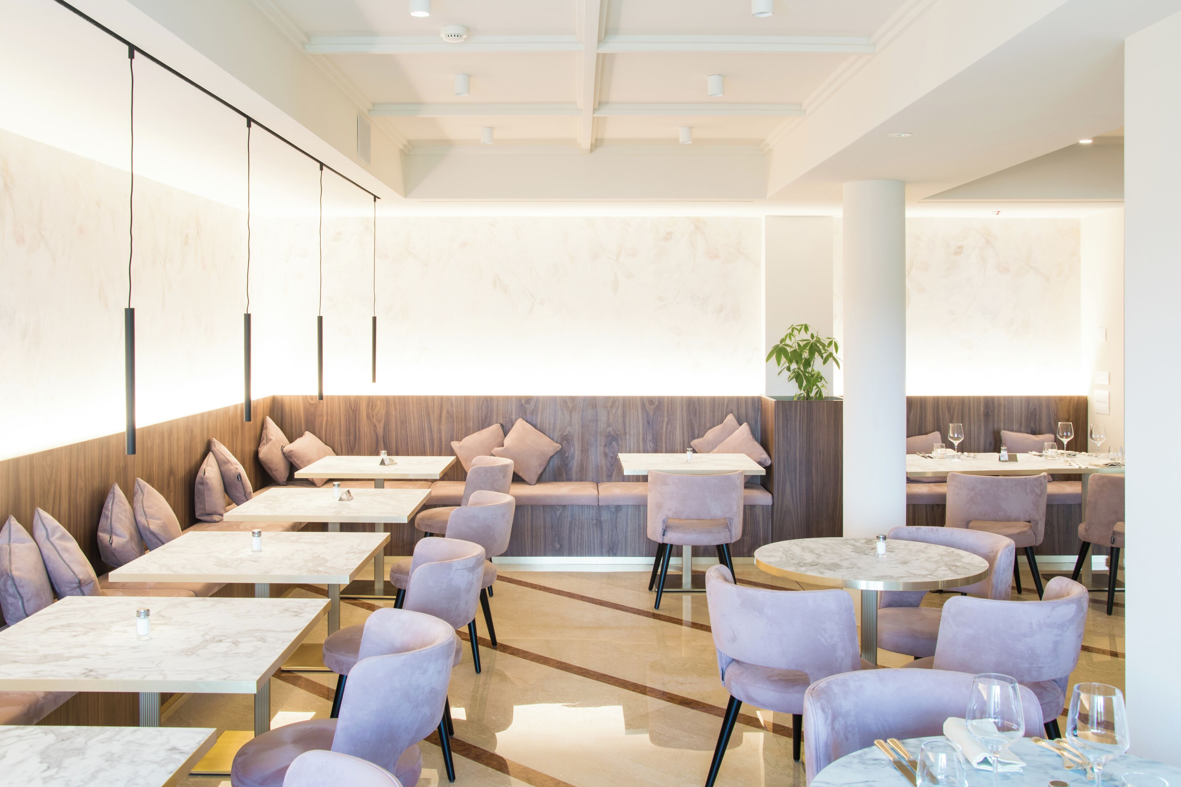 Un restaurant moderne avec des tables surmontées de marbre et des chaises rembourrées, agrémenté d’un mur d’accent en bois et d’une grande plante en pot.