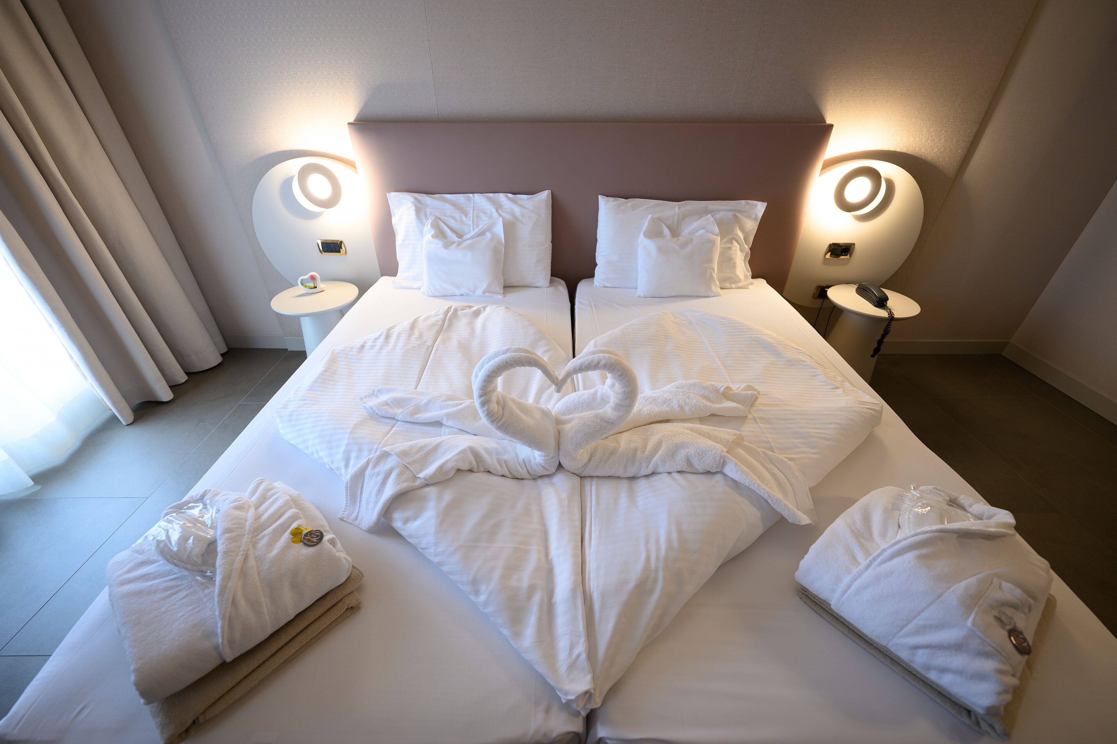 Ein Doppelbett mit einer herzförmigen Handtuchgestaltung, zwei Nachttische mit Lampen und ein Fenster mit Vorhängen.