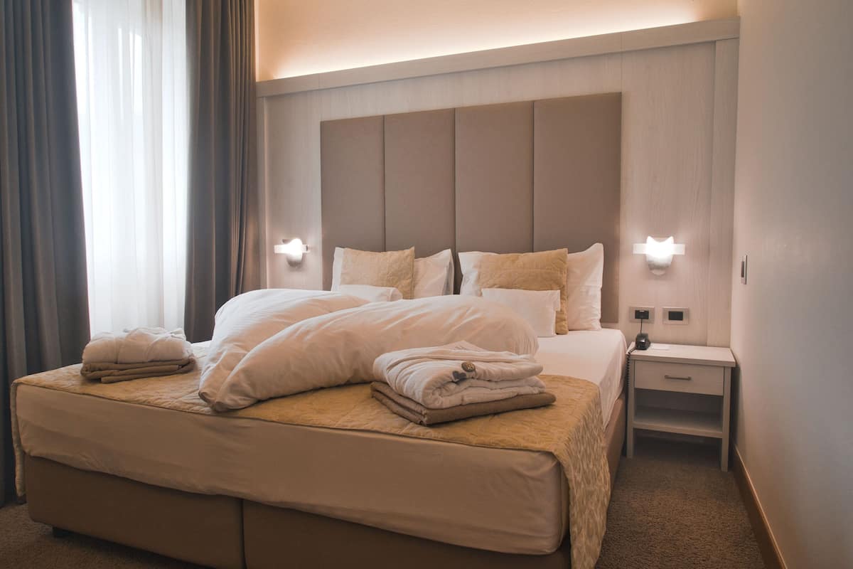 Una cama bien hecha con almohadas y un edredón doblado, una mesita de noche con una lámpara y un aplique de pared.