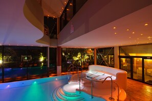 Un espace piscine intérieur moderne, doté d’un design aux lignes courbes, d’un coin salon avec un canapé blanc et d’une vue sur l’extérieur la nuit.