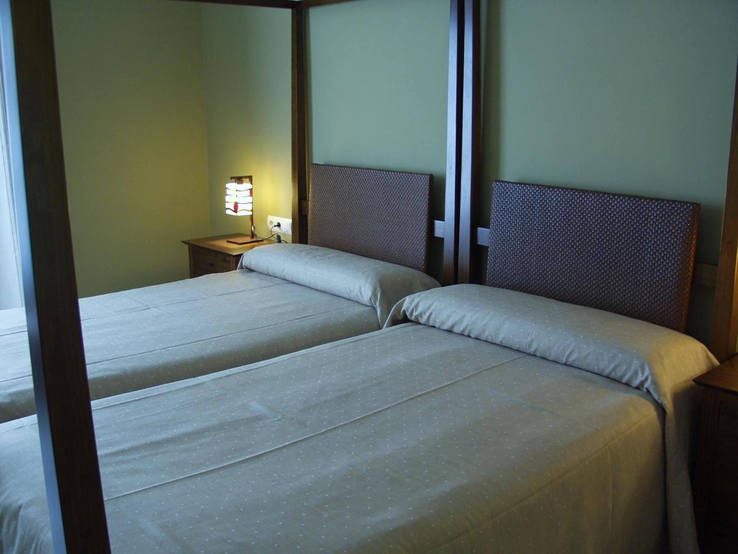 Una habitación con dos camas individuales, una mesita de noche con lámpara y un cabecero de madera.
