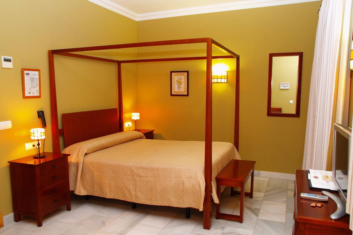 A four-poster bed with a wooden frame, a bedside table with a lamp, a mirror, and a small desk with a remote control.