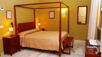 A four-poster bed with a wooden frame, a bedside table with a lamp, a mirror, and a small desk with a remote control.