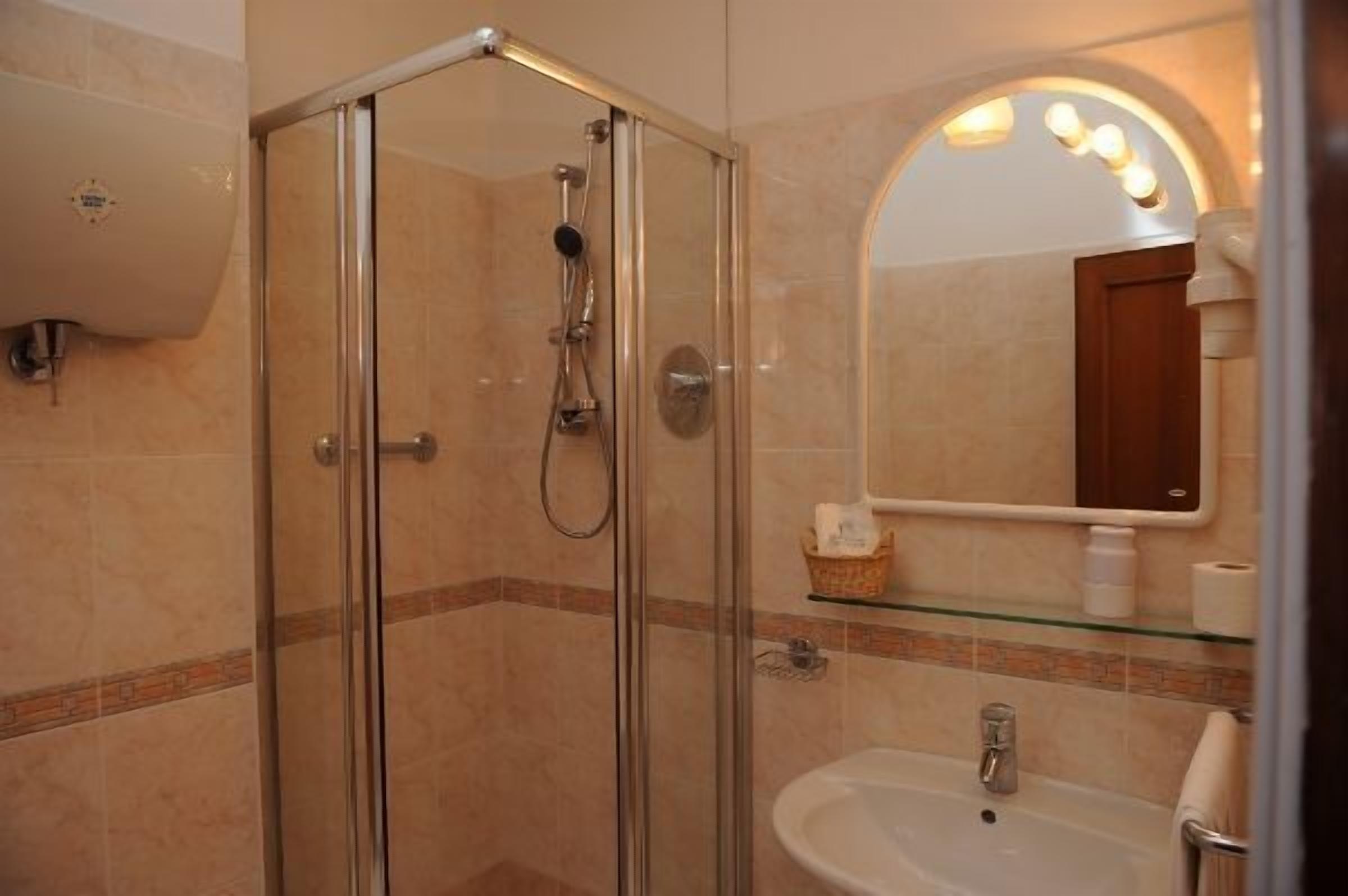 Doccia vetrata con soffione a mano, ripiano in vetro con articoli da toilette e specchio illuminato a parete.