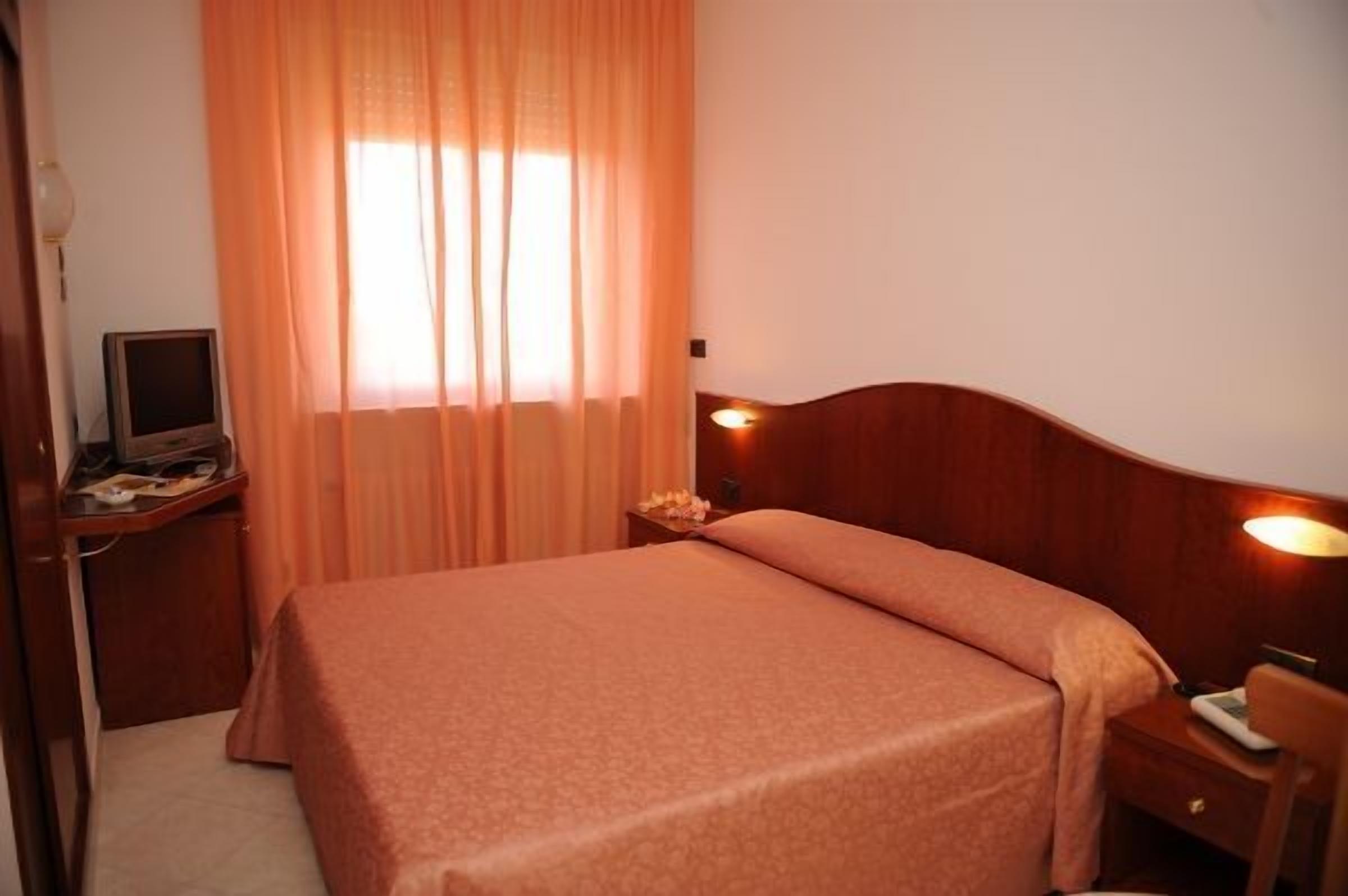 Ein Hotelzimmer mit einem Einzelbett, einem Nachttisch mit Lampe, einem Schreibtisch mit Computer und einem Fenster mit Vorhängen.