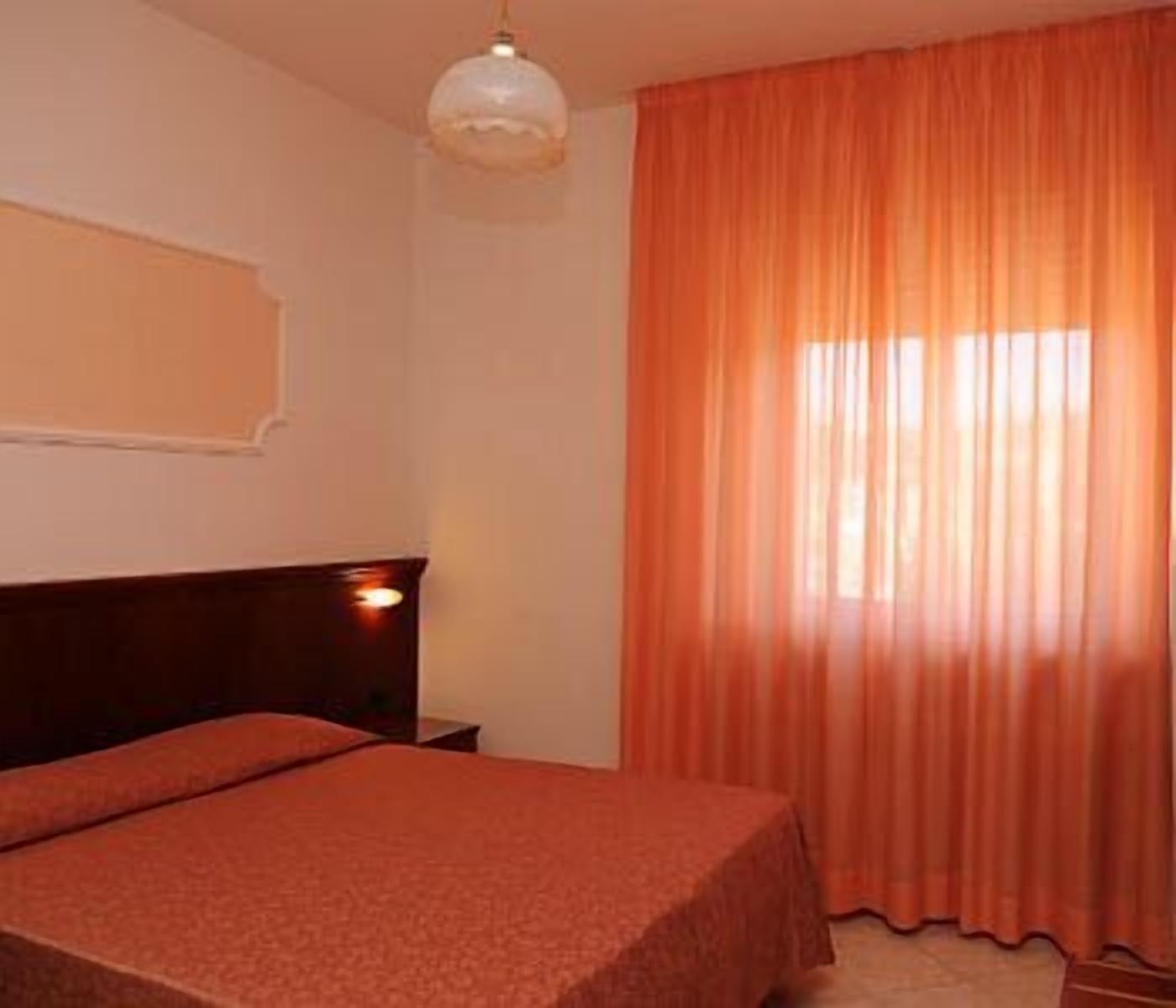 Ein Zimmer mit einem Bett, einem Nachttisch, einer Wandlampe und orangefarbenen Vorhängen.