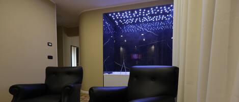 A room with two black leather armchairs, a wooden parquet floor, and a view of a dark room with string lights.