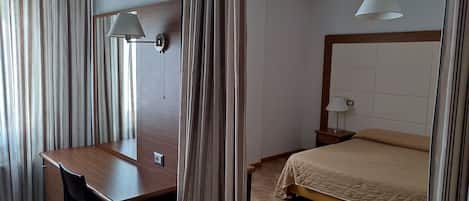 Una habitación de hotel con un escritorio de madera, una silla, una cama con colcha color beige, una mesita de noche con lámpara y una ventana con cortinas.
