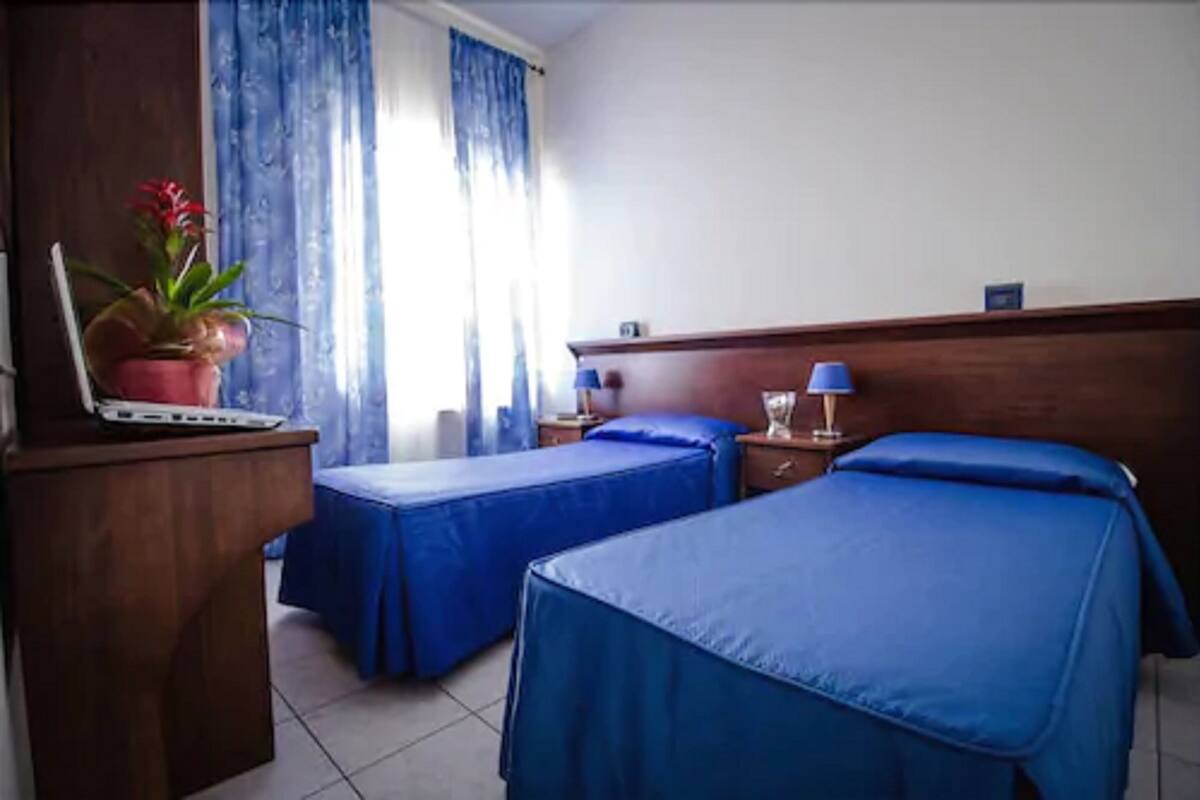 Habitación de hotel con dos camas individuales, un escritorio con una computadora portátil, una planta en maceta y una ventana con cortinas azules.