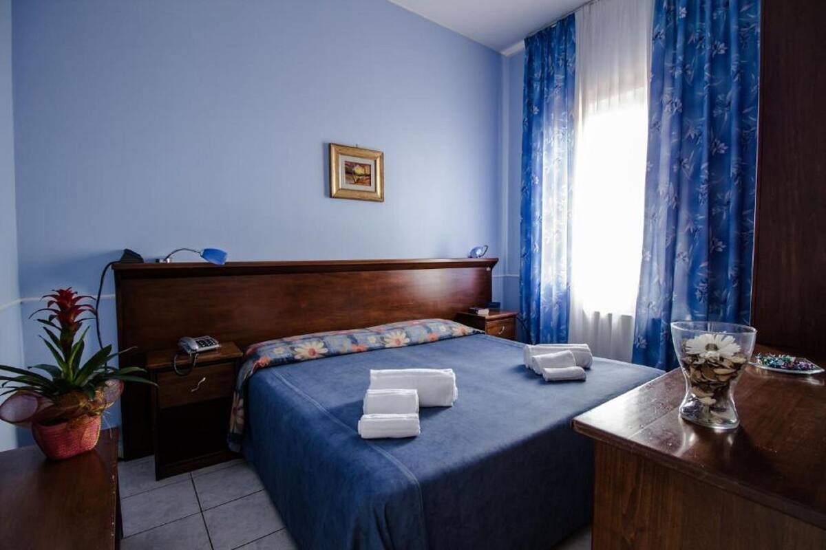 Habitación de hotel impecable con cabecera de madera, cama con ropa de cama azul, mesita de noche con lámpara, teléfono y una pequeña planta en maceta.