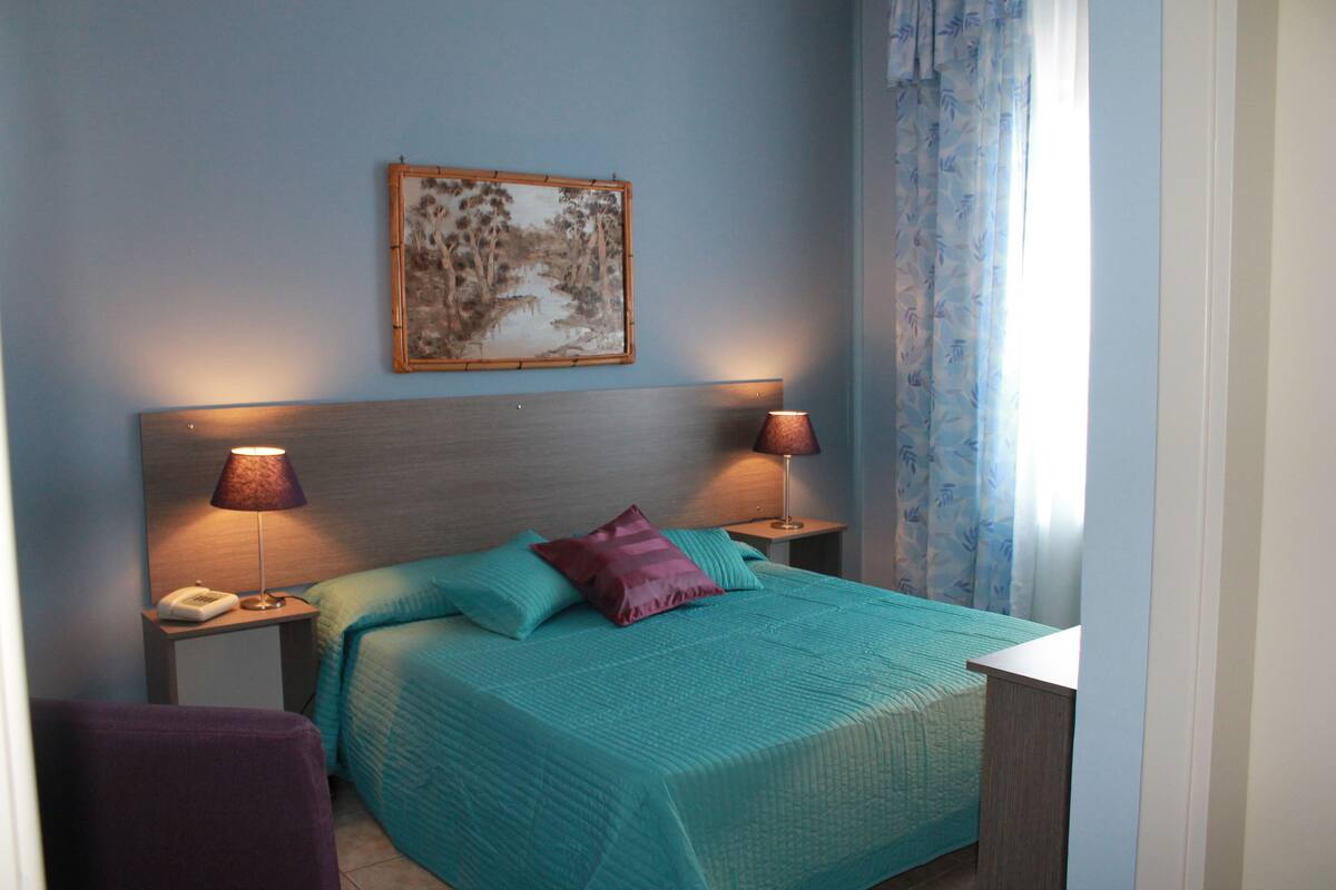 Un dormitorio con cama, dos lámparas de noche, una mesita de noche con teléfono, un cuadro en la pared y una ventana con cortinas.