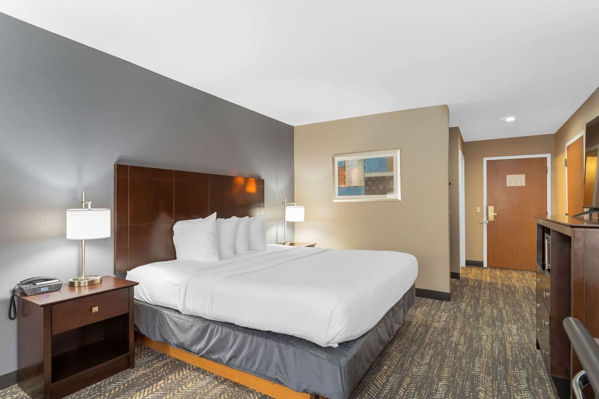 A hotel room with a bed, bedside lamps, a telephone, a desk with a lamp, a painting on the wall, and a door.