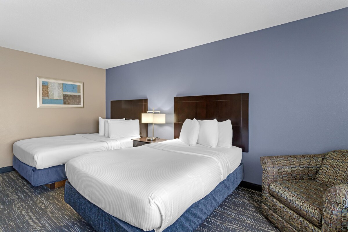 A hotel room with two beds, a nightstand with a lamp, a patterned armchair, and a framed picture on the wall.