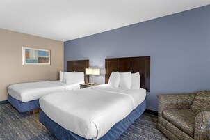 A hotel room with two beds, a nightstand with a lamp, a patterned armchair, and a framed picture on the wall.