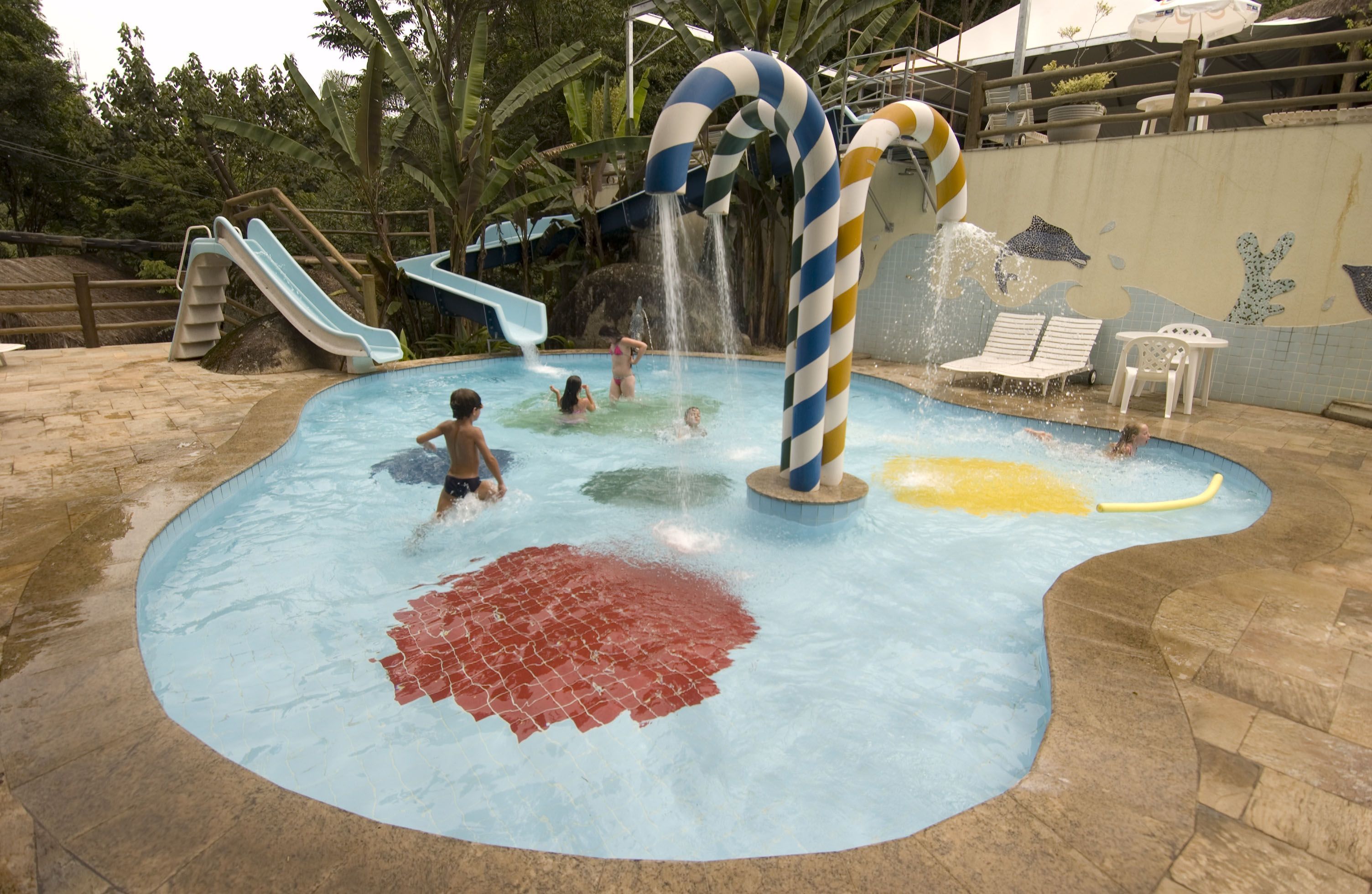Zona de juegos acuáticos con toboganes y chorros de agua, rodeada de una piscina y sillones de descanso.