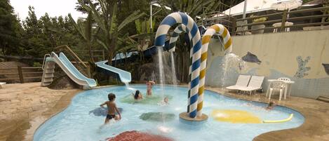 Zona de juegos acuáticos con toboganes y chorros de agua, rodeada de una piscina y sillones de descanso.