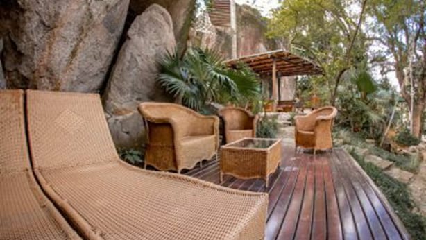 Una terraza de madera con muebles de mimbre, incluyendo un sofá y sillones, rodeada de follaje y rocas.