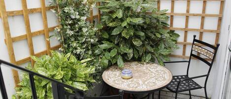 Pequeño patio al aire libre con una mesa redonda y dos sillas, rodeado de plantas en macetas y un muro de celosía de madera.