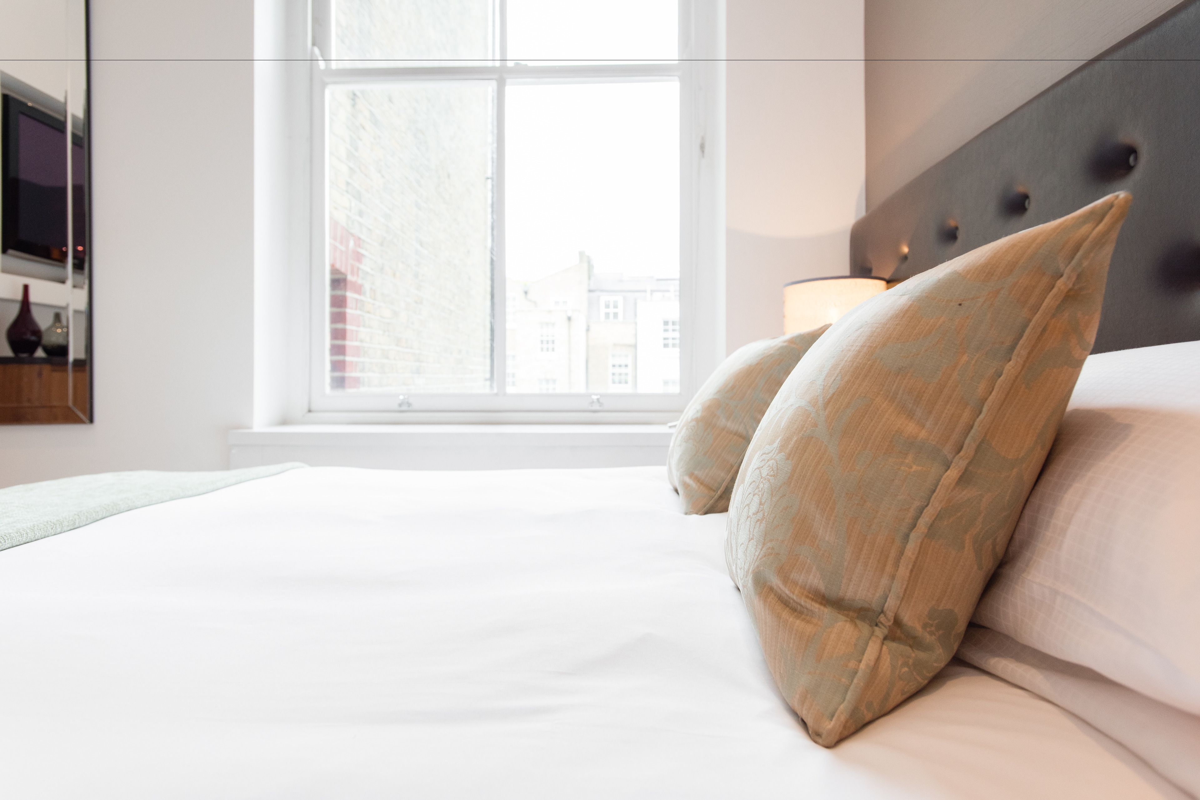 A neatly made bed with white linens and two beige pillows, a bedside table with a lamp, and a window with a view of buildings.