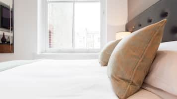 A neatly made bed with white linens and two beige pillows, a bedside table with a lamp, and a window with a view of buildings.