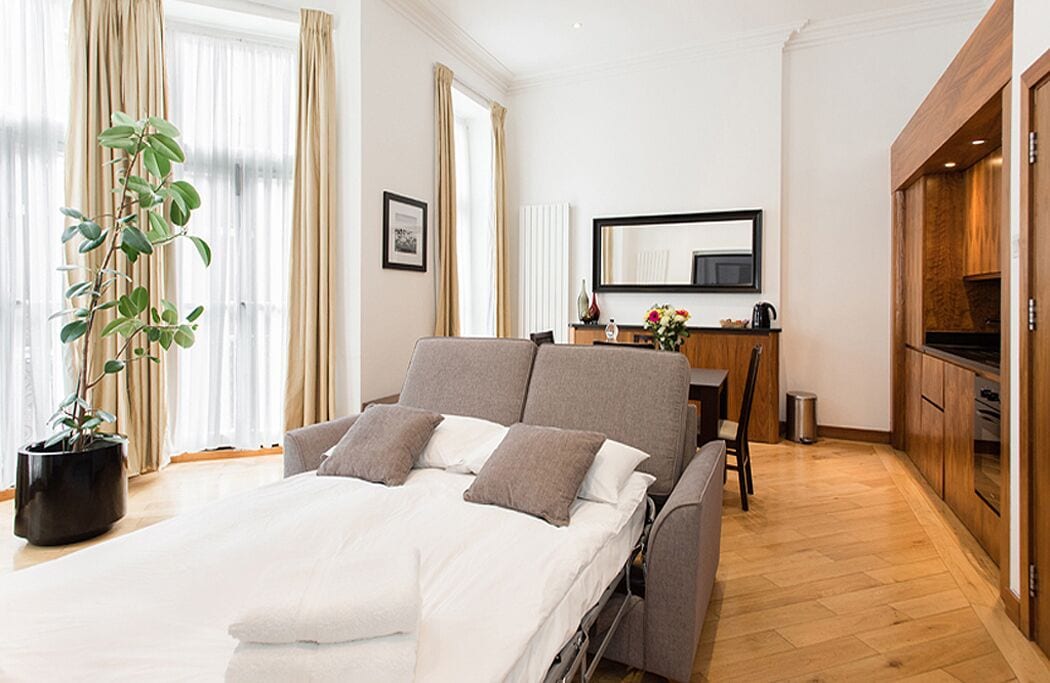 Ein modernes Hotelzimmer mit Bett, Schreibtisch, Küchenzeile und einem großen Fenster mit Vorhängen.