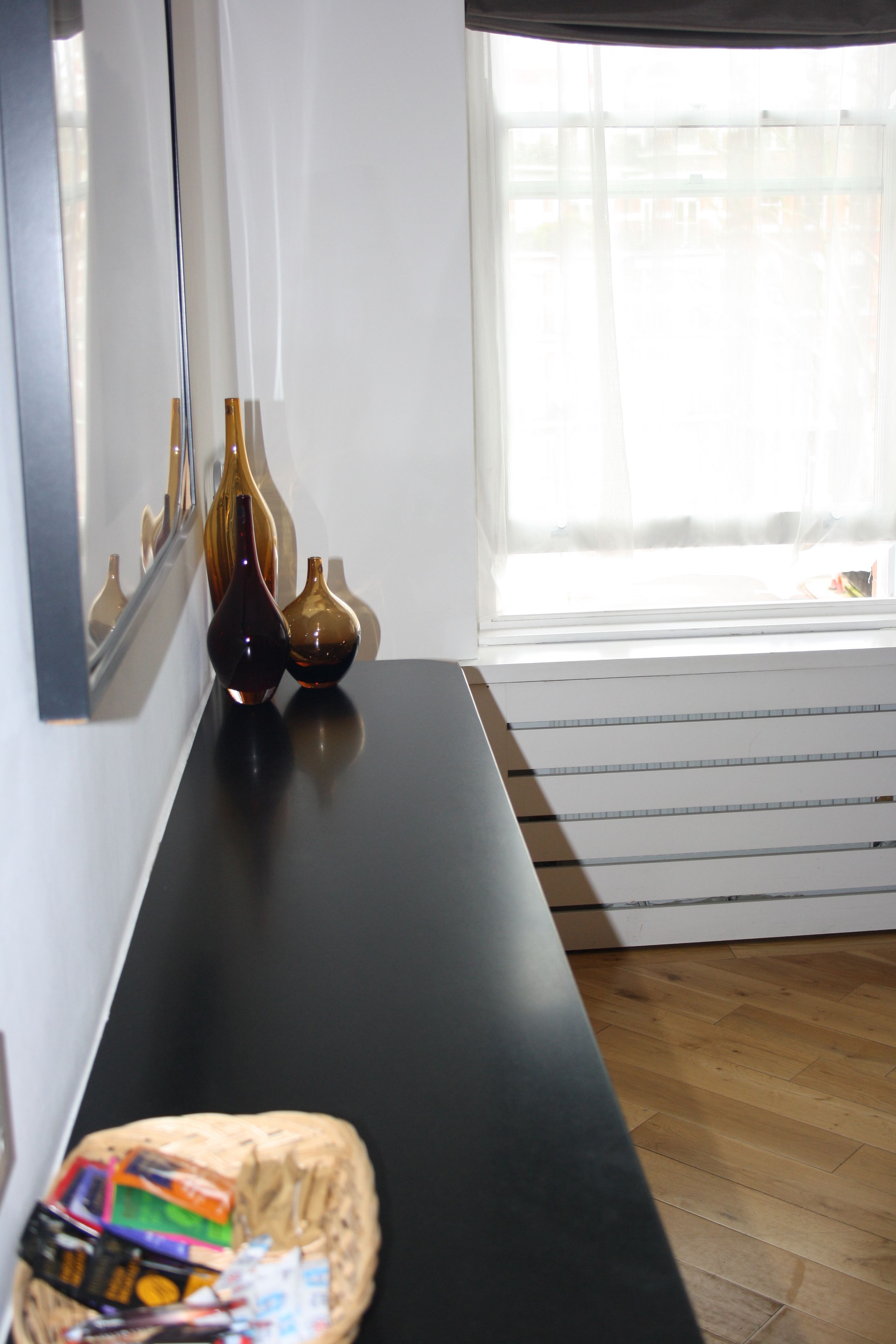 A room with a black bench, a window with sheer curtains, and a mirror reflecting vases on the wall.