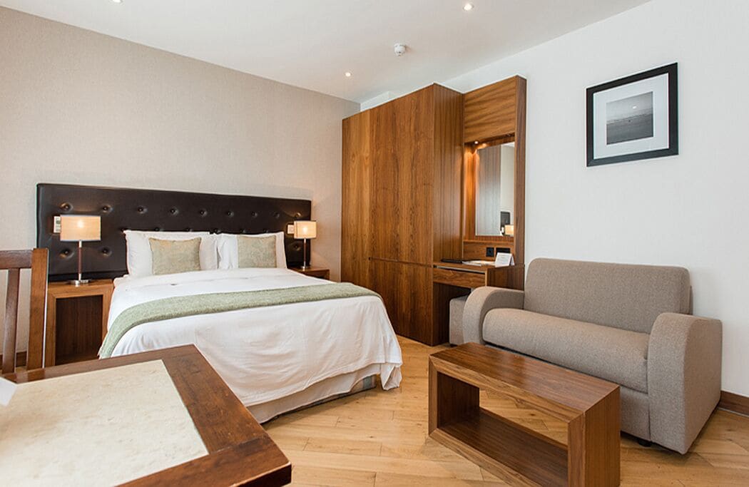 A modern hotel room with a large bed, a wooden wardrobe, a sofa, a desk, and a mirror.
