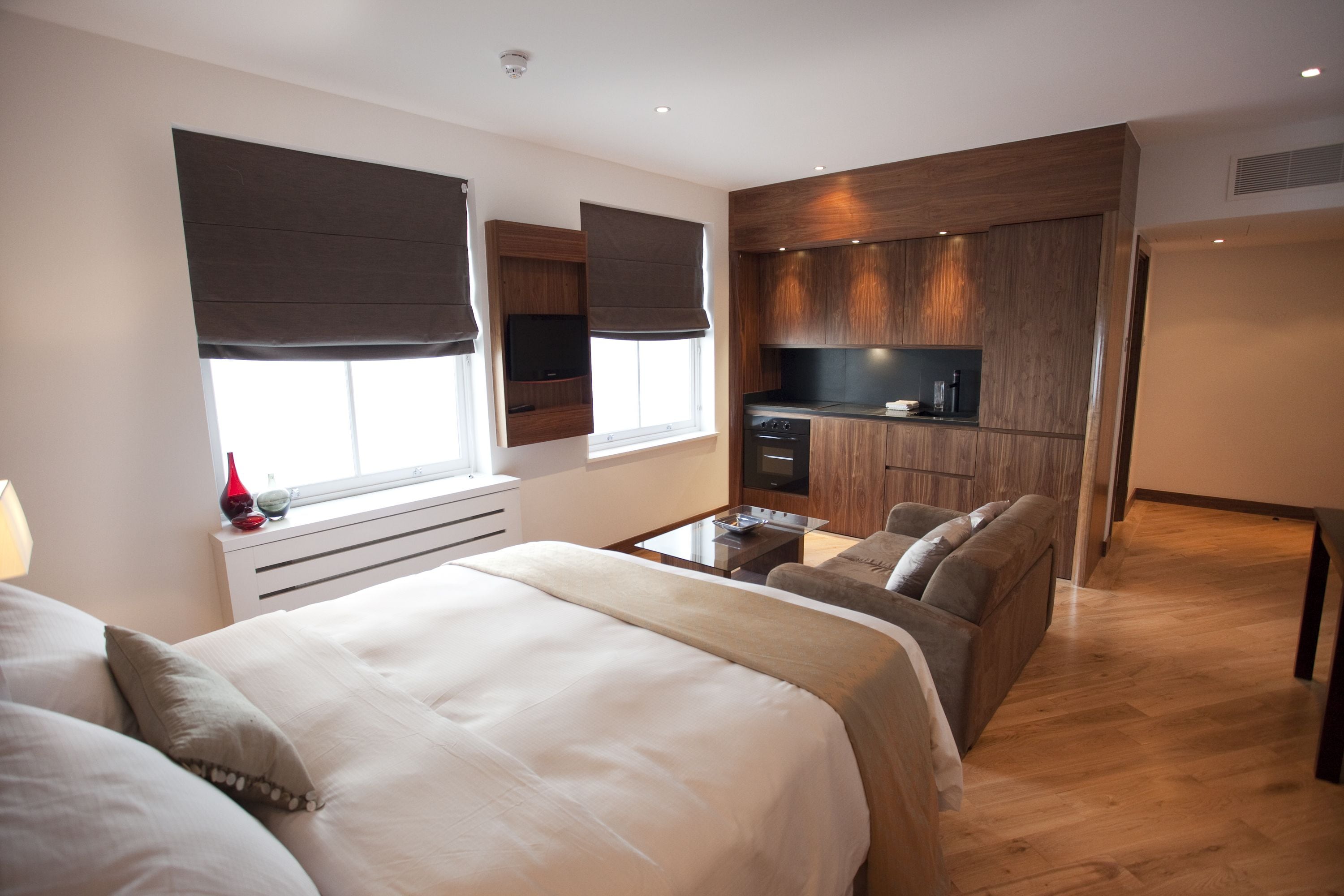 A modern bedroom with a large bed, a sofa, a small kitchen area, and a window with blinds.