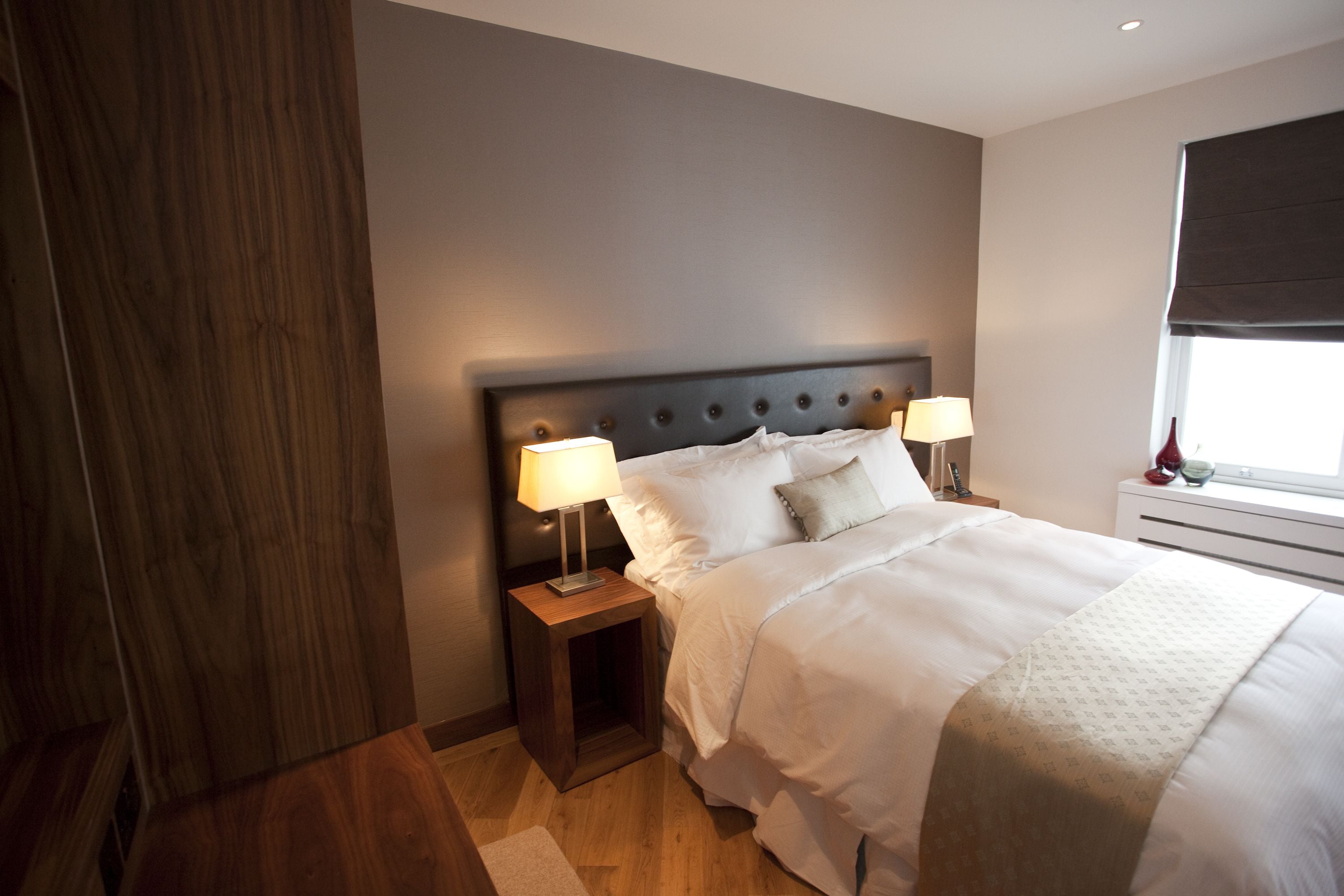 A neatly made bed with white linens, two bedside lamps, and a window with a dark blind.