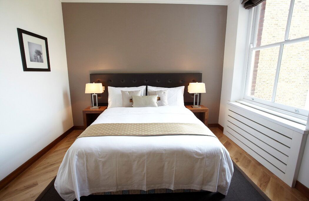 A neatly made bed with white linens, two bedside lamps, a framed picture on the wall, and a window with blinds.